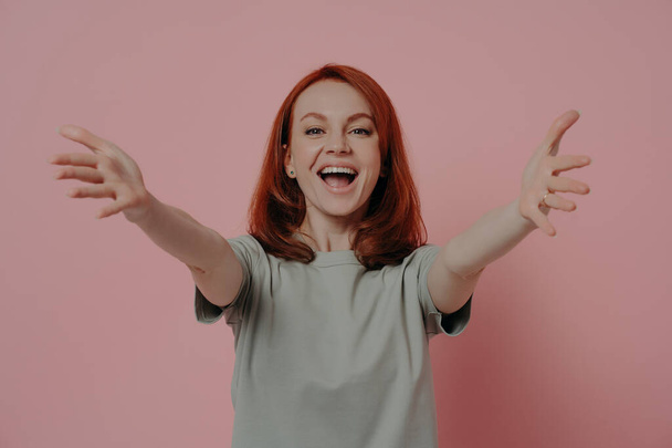 Free cuddles. Friendly happy ginger woman reaching hands forward and stretching arms for hug and smiling cheerfully while standing against pink wall, dressed in casual t-shirt - Photo, Image