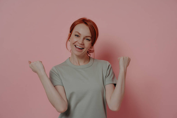 I did it! Indoor shot of overjoyed red-haired woman celebrating victory triumph, being happy, doing winner gesture and saying yes, dressed in casual t-shirt posing isolated over pink background - Valokuva, kuva