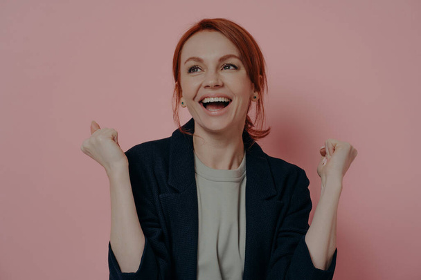 Portrait of happy excited redhead female with raised fists celebrating victory or wining and screaming with excitement and happiness, isolated on pink pastel background. Success and achievement - Zdjęcie, obraz