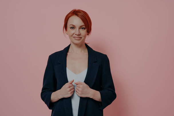 Beautiful stylish redhead female adjusting formal navy jacket and smiling at camera, expressing confidence, portrait if successful businesswoman with red hair posing isolated over pink background - Foto, imagen