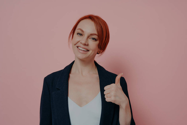 Young happy friendly ginger woman showing thumb up and looking at camera with smile, recommending something with gesture, isolated on pink background, cheerful female being satisfied with choice - Foto, Bild