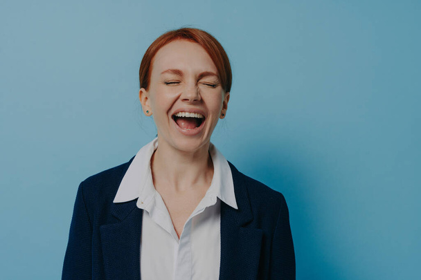 Studio shot of laughing happy woman in formal outfit with closed eyes standing isolated on blue wall, ginger female having fun, hearing funny anecdote or joke from friend. Positive emotions concept - Fotografie, Obrázek