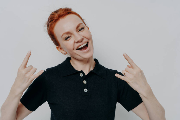 Bad girl. Cheerful carefree ginger female in casual clothes showing heavy metal rock sign while posing isolated on grey background in studio, smiling at camera rock and doing roll gesture - Photo, Image