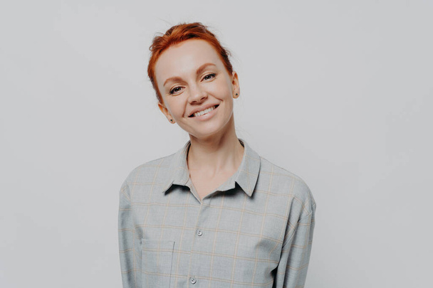 Cheerful happy beautiful red haired woman with beaming smile posing isolated on grey studio background, dressed in casual checkered shirt, expressing positivity and happiness. Face expressions concept - Photo, Image
