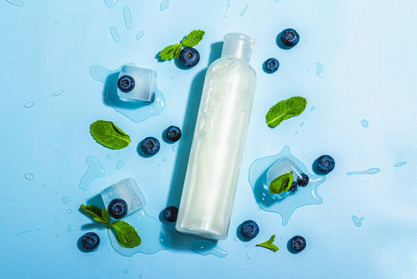 Natural skincare. Cream or lotion with organic berries. Blueberry, mint, and ice. Moisturizing and tonic cosmetics concept, flat lay, trendy hard light, dark shadow, blue background, top view - 写真・画像