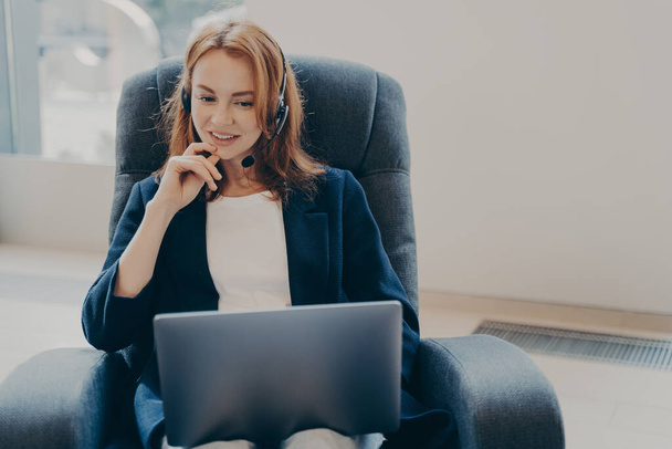Молодая привлекательная европейская женщина в наушниках сидит на кресле в домашнем офисе, принимает участие в образовательном вебинаре, улыбается женщине, используя ноутбук во время видеозвонка с клиентами, работает удаленно - Фото, изображение