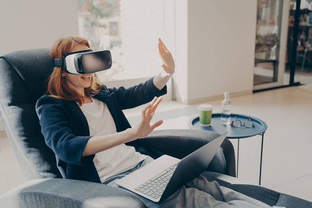 Confident young businesswoman wearing VR headset glasses and laptop sitting in cozy armchair in office lobby during her coffee break, raised hands up in air mimicking touch gestures in virtual reality - 写真・画像