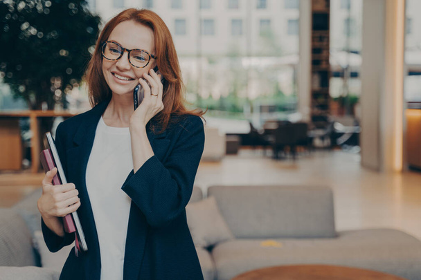 Портрет привлекательной позитивной европейской бизнес-леди в элегантном стильном наряде с мобильным разговором, деловым звонком, разговором на смартфоне в ожидании клиента или партнера в помещении - Фото, изображение