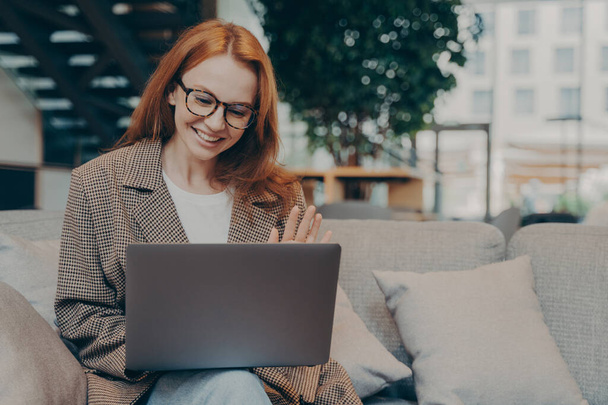 Onnellinen punatukkainen liiketoiminnan nainen optisissa silmälaseissa vilkuttaa kannettavan tietokoneen webbikamera istuessaan sohvalla ja ottaa web-konferenssi asiakkaan kanssa, harjoittaa videopuhelu kollegoiden kanssa työskennellessään etänä - Valokuva, kuva