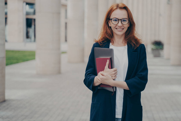 Outdoor portrait of young confident beautiful ginger woman in formal clothes holding laptop and note book and smiling at camera, waiting for business meeting, wearing spectacles, going home after work - Foto, imagen