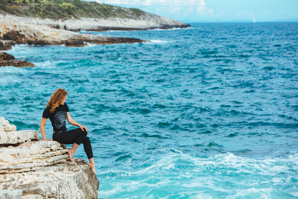Młoda szczupła kobieta siedząca na krawędzi klifu i patrząca na morze. przestrzeń kopiowania - Zdjęcie, obraz