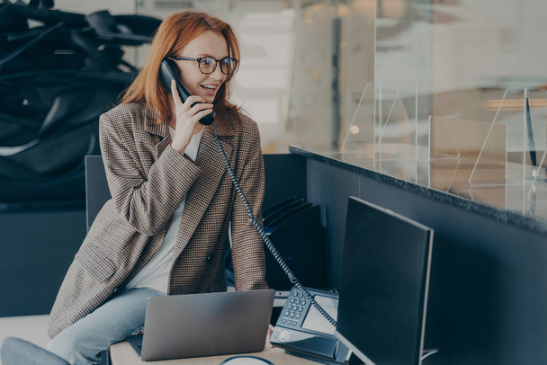 オフィスで働いている眼鏡の女性は、カジュアルな服を着て、彼女の机の上に座って電話で話をしながら、現代的で快適なオフィス環境、背景がぼやけている。仕事のコンセプトで幸せな従業員 - 写真・画像
