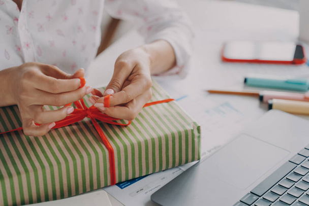 Cropped shot of woman online store business owner packing package gift box for customer during Christmas winter holidays, female tying red bow on xmas present, preparing parcel on table - Photo, Image