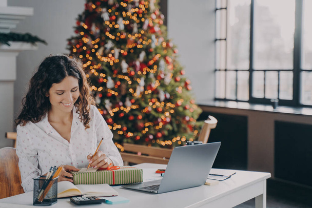 Fiatal elégedett európai nő irodai dolgozó írásban karácsonyi levél, üdvözlő kártya ünnepi kívánságait, miközben ül a munkahelyén laptoppal és távolról dolgozik otthonról a téli szünetben - Fotó, kép