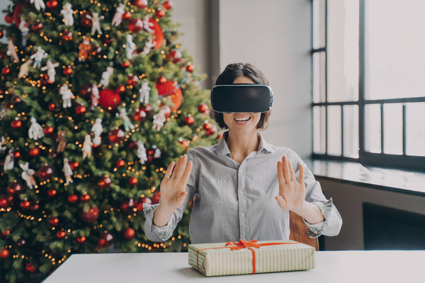 Gioiosa donna ispanica in VR auricolare seduta alla scrivania con Natale presente durante le vacanze di Natale, stupita giovane donna che si gode la realtà virtuale dei giochi 3D, albero di Natale festivo sullo sfondo - Foto, immagini