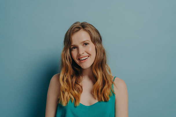 Happy young light brown-haired woman posing with head tilted, looking at camera with gentle smile opening her mouth slightly shows perfectly straight teeth isolated over blue studio background - Photo, Image