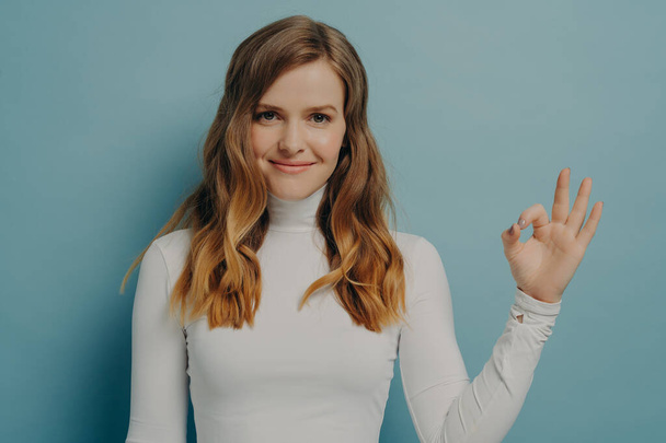 Portrait of sitisfied young europian woman showing okay gesture with fingers isolated over pastel blue stuio wall background, female demonstrating approvement. Positive body language concept - Photo, Image