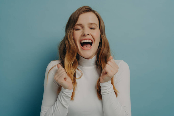 Horizontal studio portrait of young happy overjoyed woman with closed eyes clenching fists, mouth wide open with cry of delight, rejoices success, posing over blue background. Triumph concept - 写真・画像