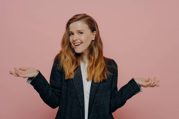 Happy attractive young woman with thick medium length ginger hair wearing dark formal jacket and white top raising hands to sides, indoor isolated on pink background. Body language concept - Photo, Image