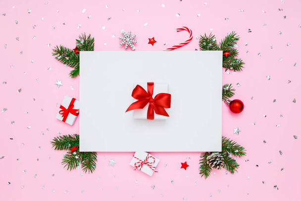 Різдвяний, новорічний святковий розклад. Біла подарункова коробка з червоним бантом в рамці гілок натуральної ялинки та прикраси на білому та пастельно-рожевому пласкому фоні
 - Фото, зображення