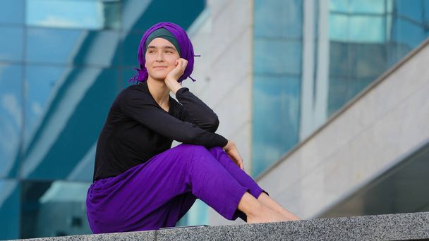 Hermosa tranquila chica islámica musulmana mujer descalza dama femenina lleva hijab púrpura se sienta en la ciudad en el edificio de la calle de fondo mirando a su alrededor descansando contemplando pensamiento inspiración soñando - Foto, Imagen