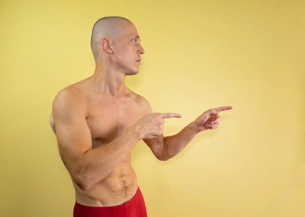 Young naked bald man in red shorts pointing with fingers on a yellow isolated background. Мужской портрет. тридцатилетний мужчина - Фото, изображение