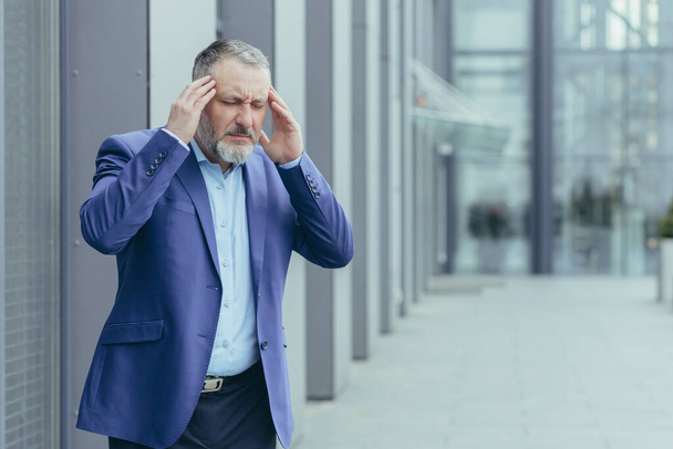 Senior gray haired businessman sick outside office building, man holding hands on head having severe migraine and headache - Φωτογραφία, εικόνα