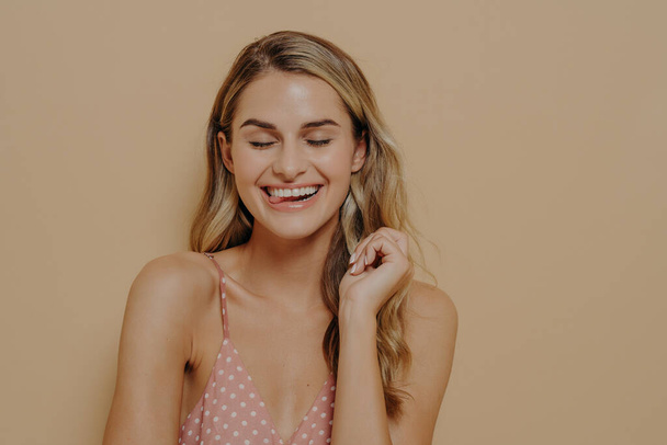 Человеческие выражения лица. Радостная веселая молодая красивая женщина с светлыми волнистыми волосами в горошек розового летнего платья смеется над шуткой, широко улыбаясь с закрытыми глазами. Концепция позитивных эмоций - Фото, изображение