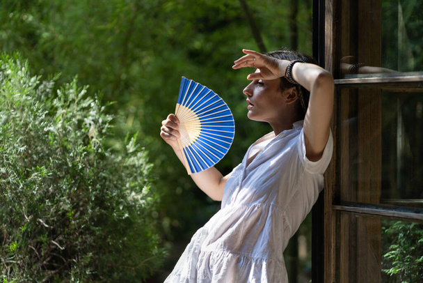 Young woman suffering heatstroke outdoors using fan to get fresh air. Unhappy girl feel bad of hot temperature outside. Frustrated female tired of summer season heat in fatigue touch forehead sweating - Zdjęcie, obraz