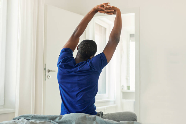 Back View Shot Of African American Young Man Stretching Arms Sitting On Bed In Modern Bedroom At Home, Wearing Blue T-Shirt. Good Morning, Weekend Concept - Foto, Imagem