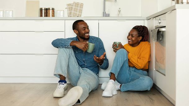 Happy african american spouses with cups drinking coffee and talking, sitting on floor in light kitchen interior, panorama with copy space. Couple relaxing together at home - Foto, imagen
