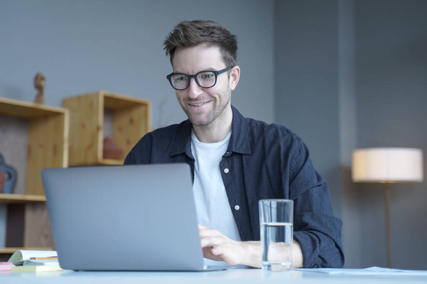 Young smiling businessman European financial consultant working remotely from home, chatting online with colleagues about details from last meeting, typing on laptop keyboard while looking at screen - Photo, Image
