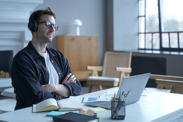 Employé de banque allemand positif assis sur le lieu de travail au bureau à domicile moderne dans des écouteurs sans fil tout en utilisant un ordinateur portable travaillant à distance en ligne, regardant l'écran écouter des cours d'enseignement à distance - Photo, image