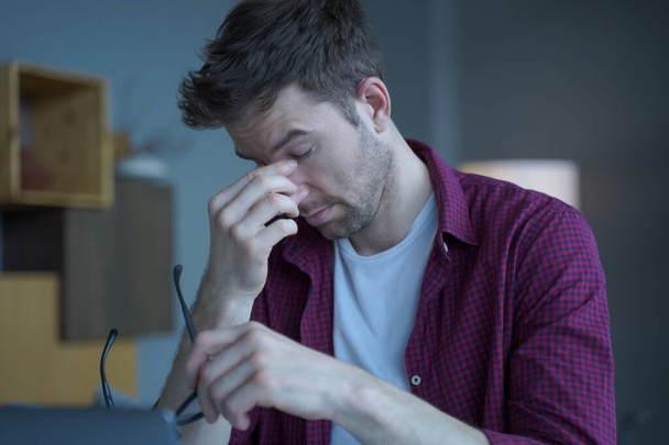 Frustrated male Austrian entrepreneur tired of remote work online sitting at modern home office, overworked man removing glasses massages bridge of his nose suffering from eye pressure and fatigue - Фото, изображение