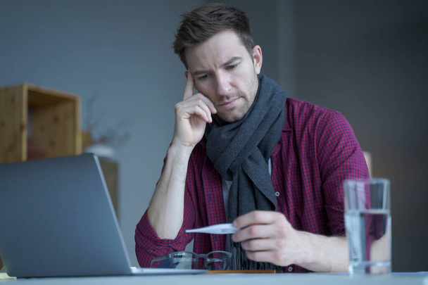 Unhealthy looking male freelancer feeling unwell and cold, wearing warm knitted scarf around neck, looking on digital thermometer in his hand checking temperature level while working remotely online - Foto, Imagem