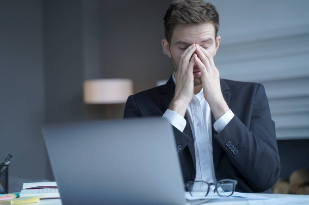Przepracowany przedsiębiorca siedzi w domu, zdejmując okulary, masując sobie nos z zamkniętymi oczami, wyczerpany człowiek cierpiący na silny ból głowy i zmęczenie. Problemy zdrowotne w pracy - Zdjęcie, obraz