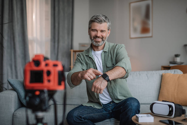 Cheerful retired european man with beard makes overview of devices and shows smart watch in living room interior. Good offer, ad and shooting video for blog at home, lifestyle, work remote and sale - Foto, Imagem