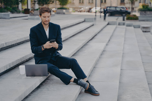 Młody pewny siebie miejski biznes profesjonalny człowiek w stylowym garniturze za pomocą smartfona podczas siedzenia na schodach na zewnątrz z laptopem. Biznesmen trzymający telefon komórkowy za pomocą aplikacji, wpisując wiadomości lub pilnej wiadomości e-mail - Zdjęcie, obraz