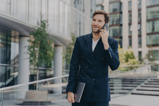 Handsome unshaven male entrepreneur communicates distantly with colleagues or partner uses modern technologies dressed in formal clothes poses outdoor near office building solves companies problems - Photo, Image
