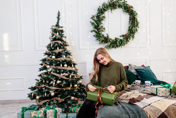 Eine junge Frau öffnet Weihnachtsgeschenke in Neujahrsatmosphäre und freut sich. Die Frau steht in einem hellen, gemütlichen Schlafzimmer, schön weihnachtlich dekoriert. - Foto, Bild