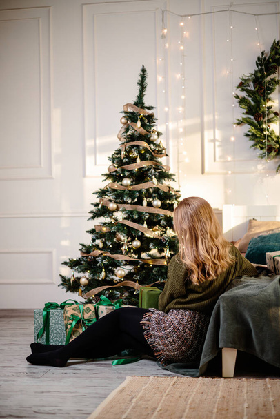 A beautiful blonde woman sits near a Christmas tree in a New Year's atmosphere and rejoices. The woman is in a bright, comfortable bedroom, beautifully decorated for Christmas. - Fotoğraf, Görsel