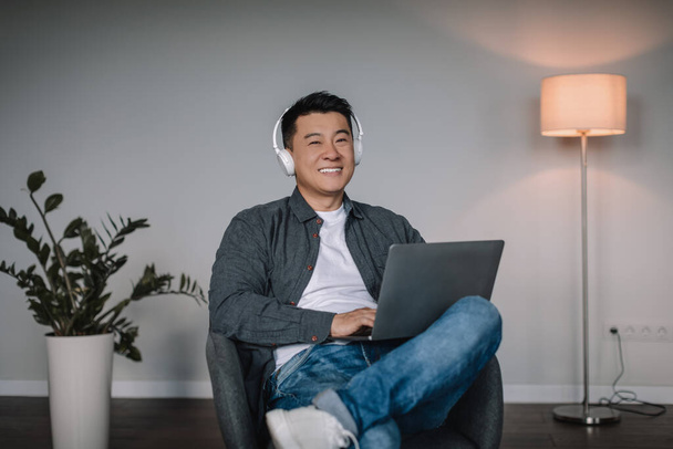 Усміхнений дорослий азіатський чоловік у бездротових навушниках насолоджується улюбленою музикою, друкує на ноутбуці, дивиться відео, сидить у кріслі в мінімалістичному інтер'єрі кімнати. Робота з пристроєм наодинці вдома, онлайн дзвінок, бізнес
 - Фото, зображення