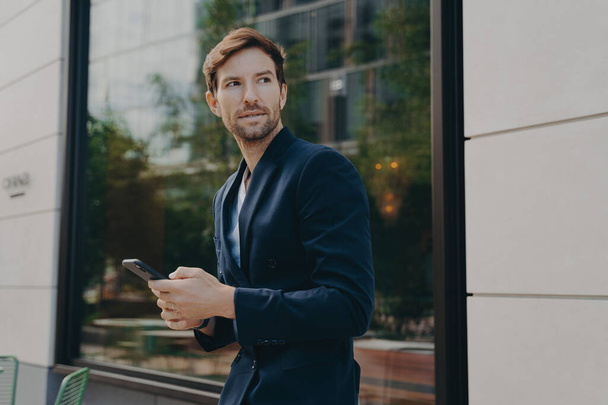 Молодий бізнесмен в темному костюмі тримає смартфон, думаючи про те, як відповісти партнеру, дивлячись далеко, стоячи поруч з офісною будівлею, вибірковий акцент на людину з мобільним телефоном
 - Фото, зображення
