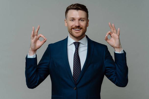 Successful bearded businessman shows ok sign with both hands dressed in formal clothes smiles pleasantly expresses approval recommends something isolated over grey background. Everything under control - Foto, Imagen
