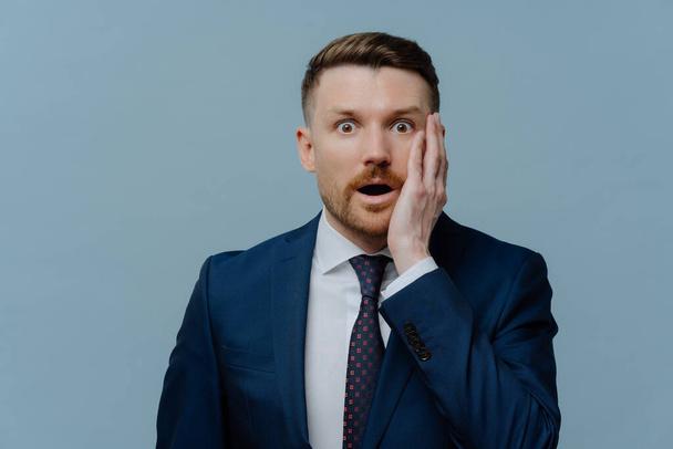 Stunned businessman in formal wear getting bad unpleasant news about his business, looking at camera with open mouth and shocked face expression while standing against light steel blue background - Photo, Image