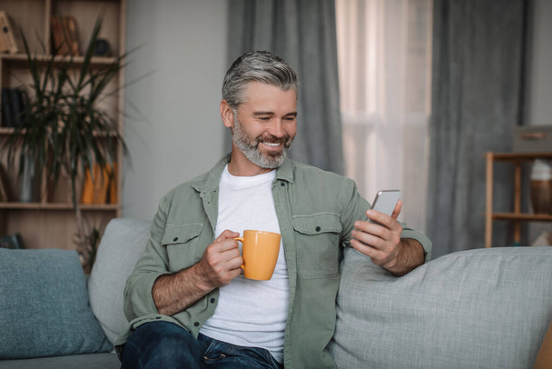 Glad retired european man with beard chatting on smartphone, enjoying drink and chat in social networks in living room interior. Ad, offer and new app at home alone in spare time due covid-19 outbreak - Фото, изображение