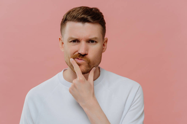 Photo of serious man holds chin purses lips makes decision with doubtful expression focused displeased at camera dressed in casual white t shirt isolated over pink background. Let me thinks a bit - Fotoğraf, Görsel