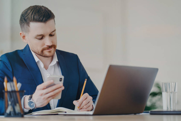 Young successful businessman with mobile phone in hand writing down important data into notepad while sitting in front of laptop at office, wearing stylish dark blue formal suit and expensive watch - Photo, Image