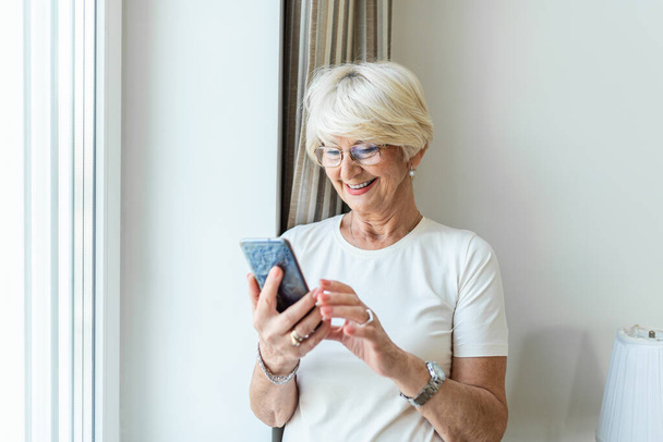 Portrait of cheerful senior woman using smartphone Photo of gray hair woman relaxing at home reading her text messages on her mobile phone with a quiet smile. Senior female texting or playing an online game on smartphone at home. - Foto, afbeelding