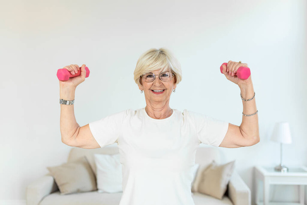 Senior woman exercising with dumbbells at home. Sporty beautiful woman exercising at home to stay fit. Elderly woman exercising at home in a living room. Fitness, workout, healthy living and diet concept. - Photo, Image